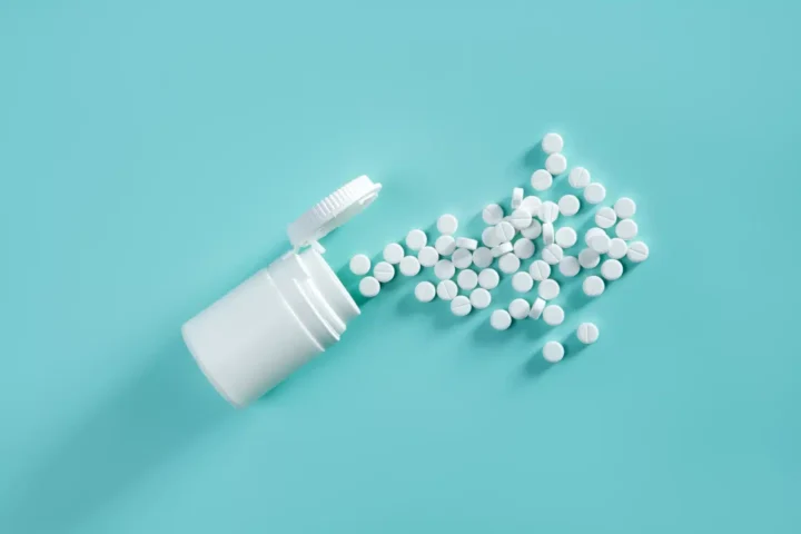 Baclofen Withdrawal Symptoms: A white bottle of pills tipped on its side with white pills spilling across a turquoise background.