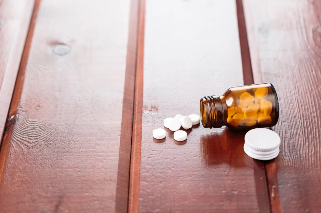 Carisoprodol Side Effects: A brown bottle containing white pills on its side with white pills spilling onto a wooden surface.