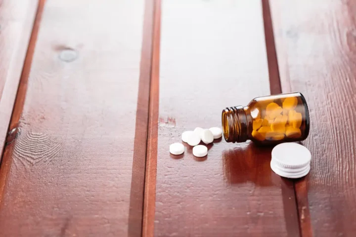 Carisoprodol Side Effects: A brown bottle containing white pills on its side with white pills spilling onto a wooden surface.
